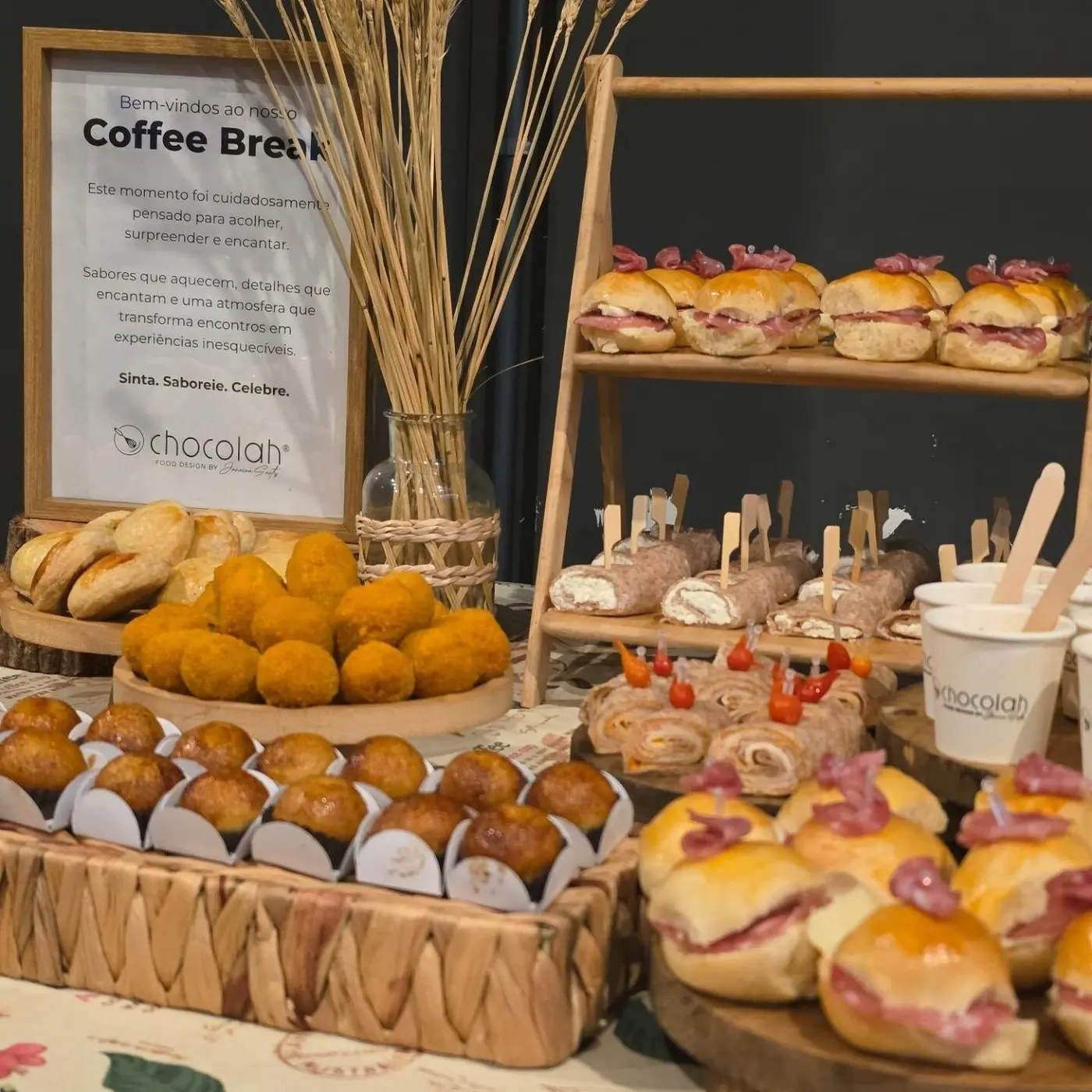 Mesa de coffee break montada pela Chocolah, com salgados, doces e placa de cardápio